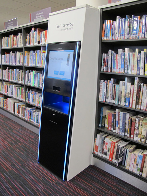 Self-service kiosk with illuminated edges beside browsing shelves at Caerphilly Library