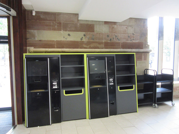 Self-service kiosks with black and lime green finishes alongside shelving units in the entrance area at Lichfield Library